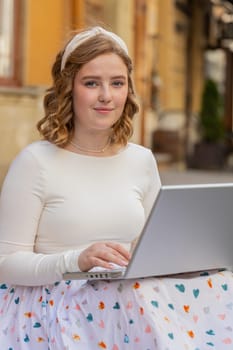 Woman freelancer sitting on city street using laptop working online distant job browsing website chatting outdoors during break. Girl looking at camera white sends messages watching movies at notebook