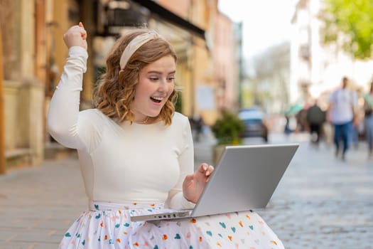 Overjoyed happy young Caucasian woman working on laptop scream in delight raise hands in triumph winner gesture celebrate success win money outdoors. Happy girl sitting on downtown street in city.