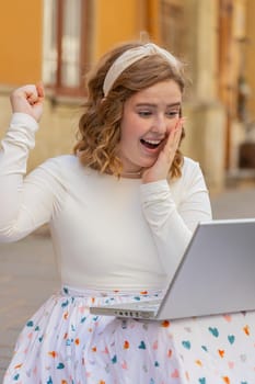 Overjoyed happy young woman working on laptop scream in delight raise hands in triumph winner gesture celebrate success win money outdoors. Happy girl sitting on downtown street in city. Vertical