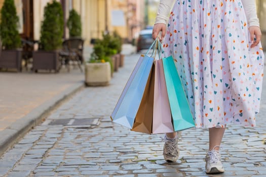 Anonymous woman shopaholic consumer after shopping sale with colorful bags with gifts in hand outdoors. Close-up of girl tourist traveler walking along the urban city street road. Town lifestyles.