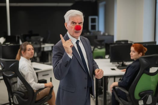 Two Caucasian women look at a serious elderly man with a clown nose in the office