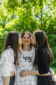 Portrait of a young Caucasian mother with two adult daughters who are kissing her on both cheeks while standing in a city park on a summer day, side view.