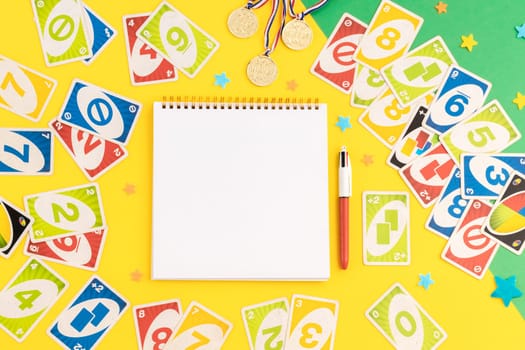 Old,dirty,colorful uno game cards,three winner medals,empty notepad,pen and felt stars lie on a green-yellow background with copy space in the center,flat lay close-up.Summer board games concept.