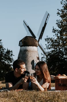One beautiful caucasian young couple lies on their stomachs on a bedspread with a wicker basket, fruits and tenderly look at each other, holding glasses of champagne in their hands in a park against the backdrop of an old windmill, close-up side view.