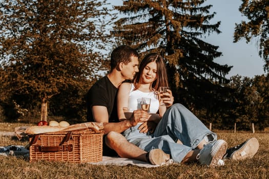 One beautiful caucasian young couple holding glasses of champagne sit hugging on a bedspread with a wicker basket and fruits in the park on a summer sunny day, close-up side view.