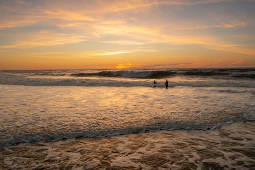 Two surfers are training to ride waves in Atlantic Ocean at sunset wearing wetsuits. Aerial view