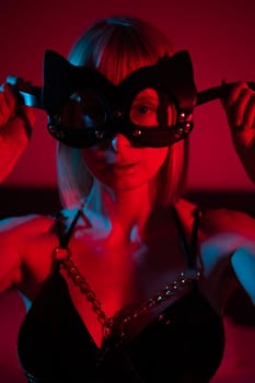 Blonde woman putting on leather mask in bedroom in red blue neon light