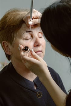 One young Caucasian brunette girl cosmetologist paints eyebrows with brown paint using a brush to a beautiful elderly woman in a home beauty salon, side view close-up.