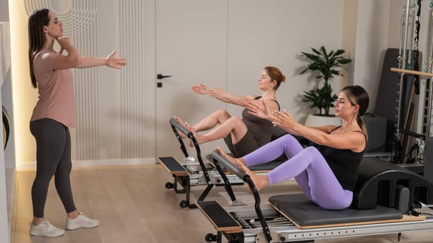 Two pregnant women are doing Pilates on a reformer. The instructor teaches prenatal yoga