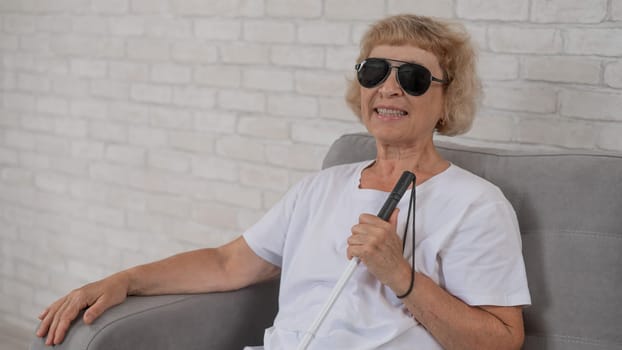 An elderly blind woman wearing sunglasses and with a tactile cane sits on the sofa