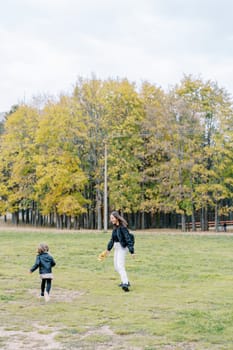 Smiling mother and little girl are running through a green meadow at the edge of an autumn forest. High quality photo