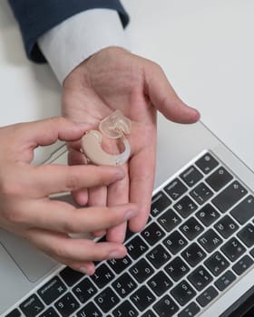 Close-up of a man's hands with a hearing aid near a laptop. Vertical photo
