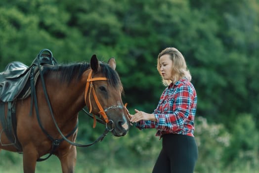 Happy blonde with horse in forest. Woman and a horse walking through the field during the day. Dressed in a plaid shirt and black leggings