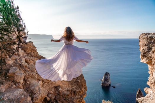 A woman in a white dress stands on a rocky cliff overlooking the ocean. The scene is serene and peaceful, with the woman's dress billowing in the wind. The ocean is calm and blue