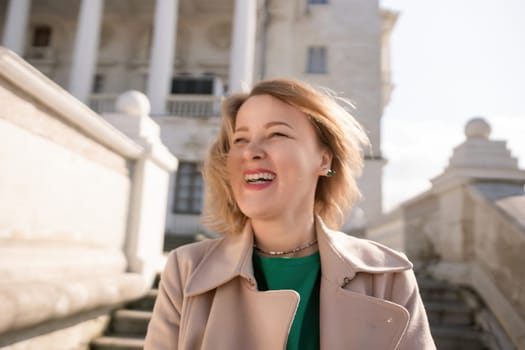 A woman with long hair is standing on a set of stairs. She is wearing a green shirt and a tan coat