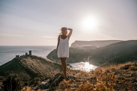 A blonde woman stands on a hill overlooking the ocean. She is wearing a white dress and she is enjoying the view