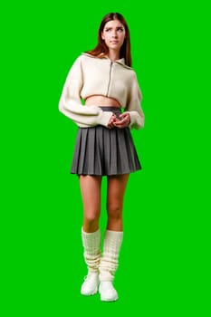 A young woman stands against a vibrant green backdrop, her gaze directed off-camera with a thoughtful expression. She wears a casual, cropped white zip-up sweater paired with a dark pleated skirt, and her long brown hair hangs straight over her shoulders.