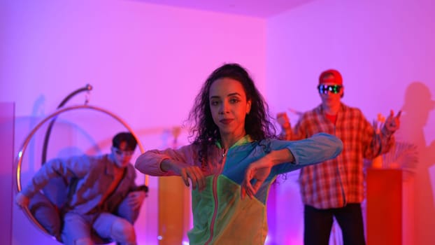 Attractive happy dancer looking at camera while smart woman moving at rhyme with neon light. Professional hispanic performer break dancing while wearing colorful cloth with diverse friend. Regalement.