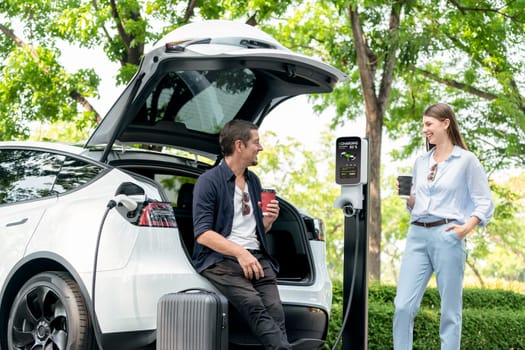 Lovely young couple drinking coffee while recharging battery for electric car during road trip travel EV car in natural forest or national park. Eco friendly travel during vacation and holiday. Exalt