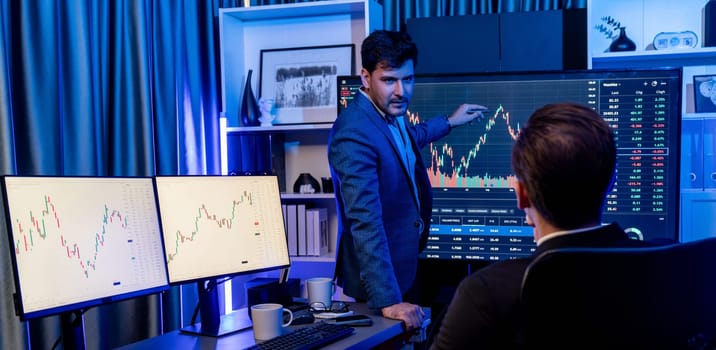 Two stock exchange traders discussing dynamic valued investment rate on monitor at night in panorama photo view. Businessman in stock market presenting economic in neon light at workplace. Sellable.