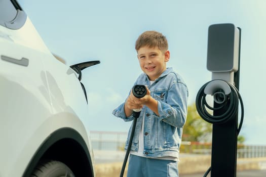 Playful little boy pointing EV charger at camera, recharging eco-friendly electric car from EV charging station. EV car travel by the seashore using clean and sustainable energy.Perpetual