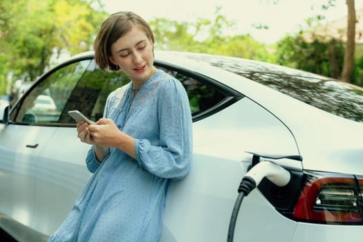 Holiday road trip vacation by the green countryside nature with beautiful young woman checking battery status from smartphone while recharging electric vehicle.Eco-friendly travel wit EV car.Perpetual