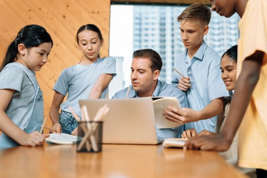 Teacher sitting and giving a lecture on lesson to children in classroom using laptop, talking to children about content in notebook. Students asking about subject instructor teach them. Edification.