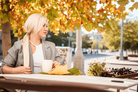 A blonde woman sits at a table with a cup of coffee and a leaf on it. The scene is set in a city with a tree in the background