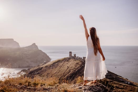 A woman in a white dress stands on a rocky hill overlooking the ocean. She is smiling and she is happy