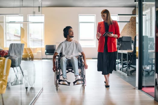 A business leader with her colleague, an African-American businessman who is a disabled person, pass by their colleagues who work in modern offices.