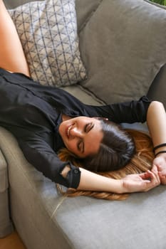 Closeup of a young model, dressed in a black shirt, lying on her back on the sofa, arms stretched out over her head, smiling with her eyes closed.