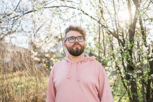 Man with beard and mustache on smiling face near sakura flowers or blooming spring tree. Soft and gentle concept. Bearded man with stylish haircut with flowers on background, close up. Hipster near branch of bloom tree