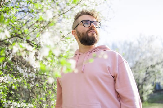 Man with beard and mustache on smiling face near sakura flowers or blooming spring tree. Soft and gentle concept. Bearded man with stylish haircut with flowers on background, close up. Hipster near branch of bloom tree