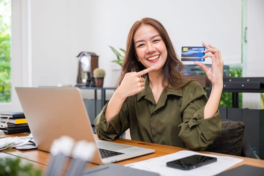 E-commerce concept. Asian young woman excited holding credit card and pointing the card with laptop computer at home office, paying shopping online, Beautiful female using laptop while sitting on desk