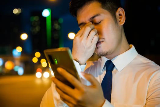 Tired businessman in suit rubbing eyes after using smartphone at night. Frustration due to eye strain serious about care concerns for eyesight street scene.