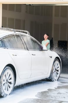 Denver, Colorado, USA-September 1, 2023-A young girl enthusiastically assists in washing the family's electric car in their suburban driveway.