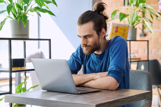 Young serious man working and studying using laptop sitting in coworking cafe. 30s handsome bearded guy looking at computer screen. Technology for leisure work business education communication people