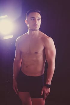 A man with a bare chest is posing against a dark backdrop, showing off his muscular physique. His head, hands, stomach, arms, shoulders, legs, and neck are visible in the flash photography