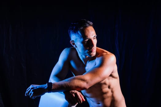 A man with a bare chest is posing against a dark backdrop, showing off his muscular physique. His head, hands, stomach, arms, shoulders, legs, and neck are visible in the flash photography