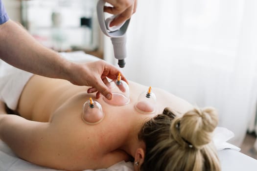 A woman is receiving a cupping treatment on her shoulder at a spa. A hand is applying cups on her skin to improve joint function and relieve tension