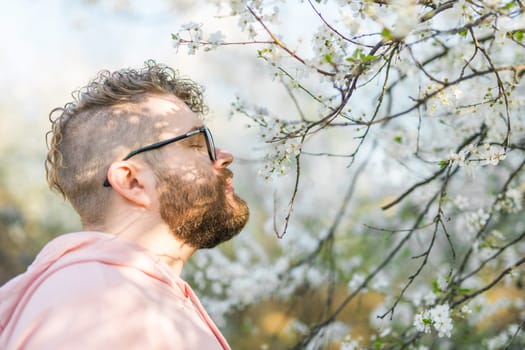 Portrait of curly millennial man inhales the fragrance of spring flowers of blooming jasmine or cherry tree. Spring time