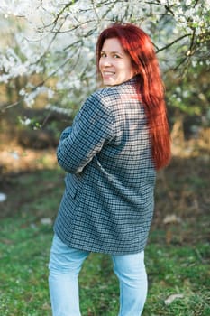 Beautiful young red hair woman near blooming spring trees alley. Youth and love, fashion and romantic and lifestyle concept