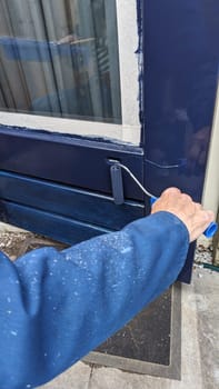 a man in blue overalls is busy painting an outside door blue with a roller, the windows are taped up