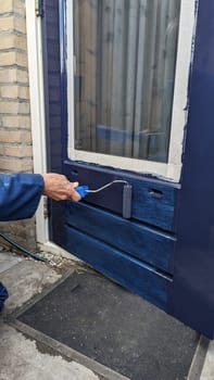 a man in blue overalls is busy painting an outside door blue with a roller, the windows are taped up