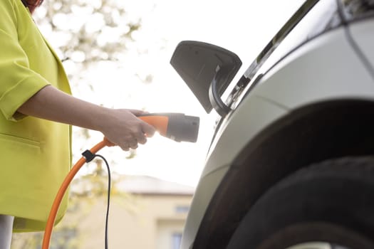 Close up shot of unrecognizable caucasian female hands opening an electric car charging socket cap and plugging in a charger. Woman charging the modern zero emission car on clean sky background.