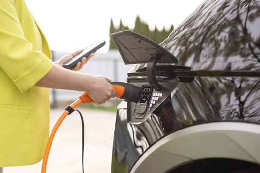 Woman connects an electric car to the charger and adjusts the process of charging the car battery using a cell phone smart phone. Girl plugs power cable to charge electric car in parking lot