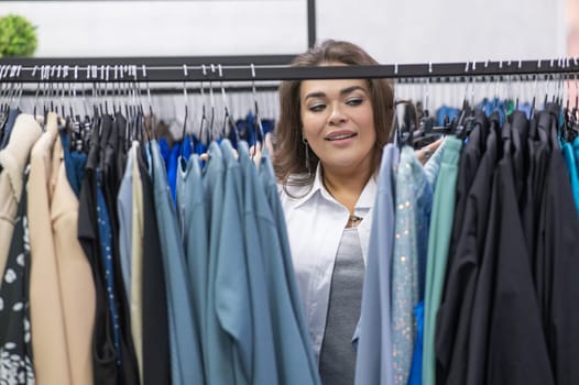 Portrait of a fat woman in a plus size store through hangers with clothes