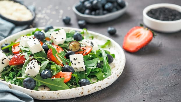 Salad with arugula, feta cheese and berries - strawberry, blueberry, in craft plate on black bacground. Summer salad idea and recipe for healthy vegetarian lunch, dinner. Copy space.