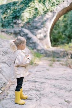 Little girl stands on a paved path in a green park and looks at the arched bridge. Back view. High quality photo