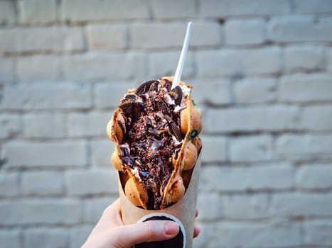 Hong Kong waffles in female hand on brick wall background. Delicious bubble waffle with chocolate biscuit cookies and chocolate syrup in woman hand. Trendy food.